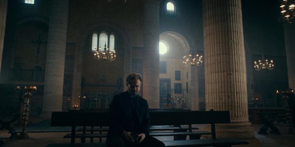 Father Vincent sits on a bench inside looking pensive.