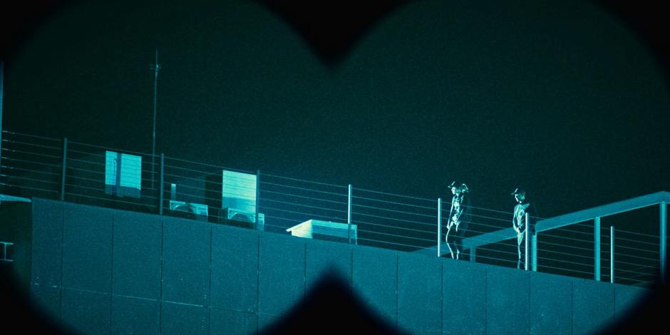 Two guards are visible on the roof through the view of some binoculars.