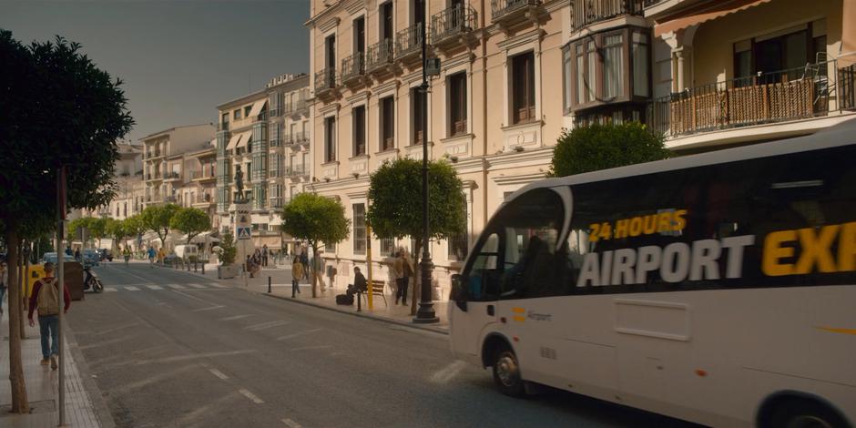 The airport bus drives down the street towards where Beatrice is waiting.