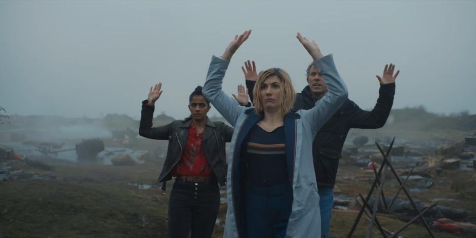 Yaz, the Doctor, and Dan raise their hands above their heads when approached by a figure through the fog.