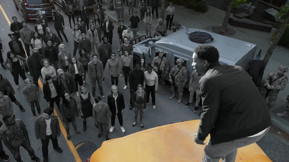 A crowd of grey people listen to Orlando's speech from atop a school bus.