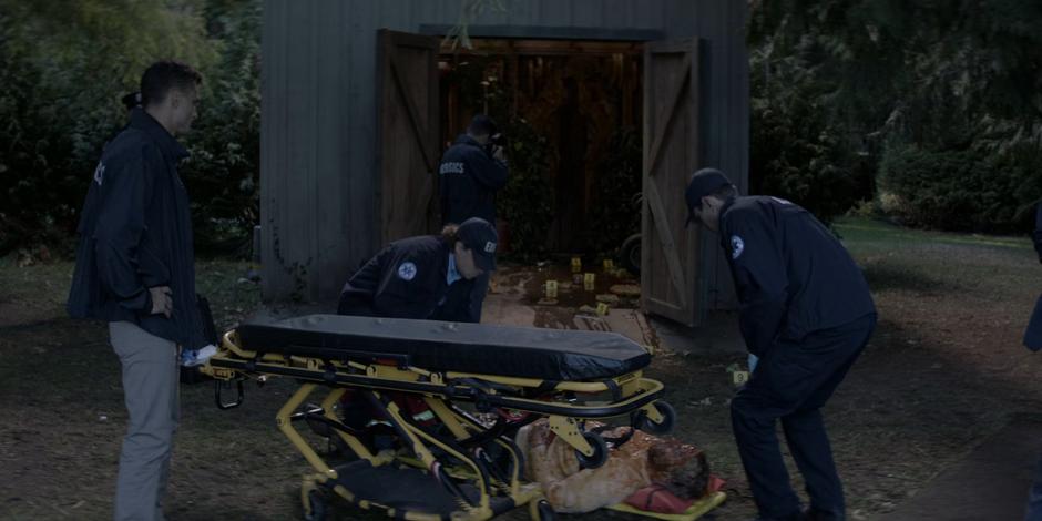 EMTs load the man onto a stretcher while forensics techs photograph the shed.