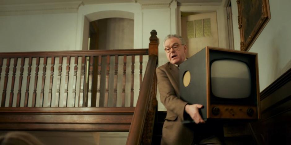 Professor Jericho carries a TV down the stairs in the main hall.