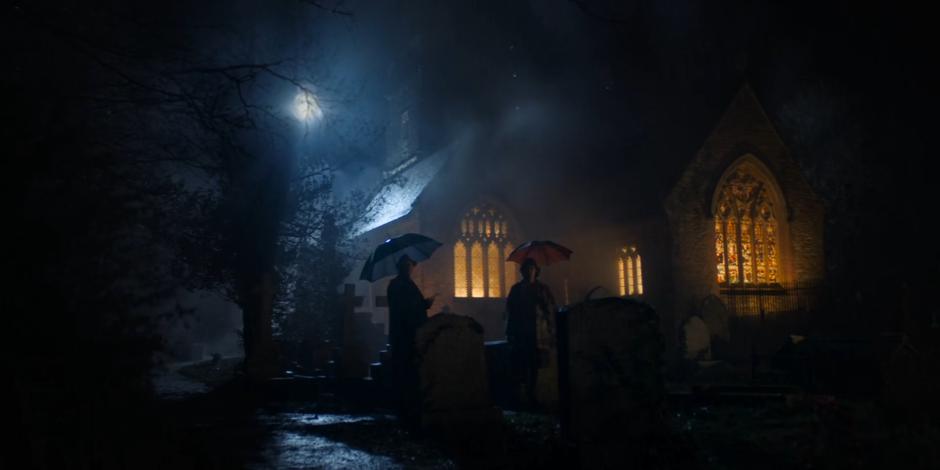 Reverend Shaw joins Mrs. Hayward in the graveyard under umbrellas.