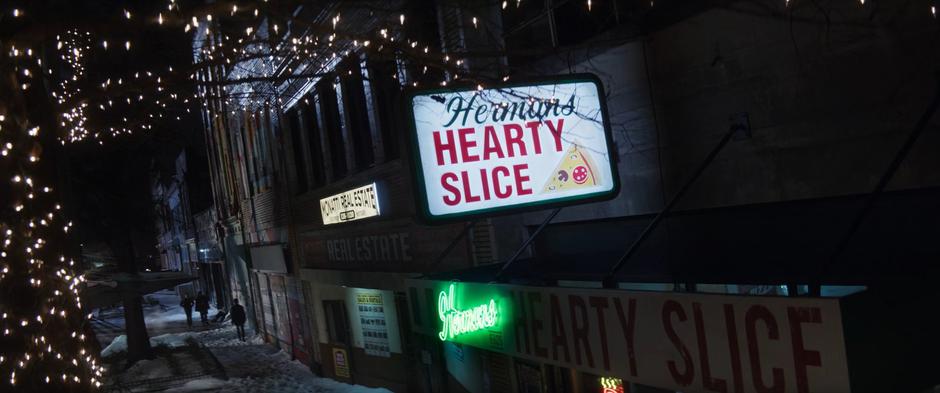 A sign for Herman's Hearty Slice hangs in the foreground as Kate and Clint walk down the sidewalk in the distance.