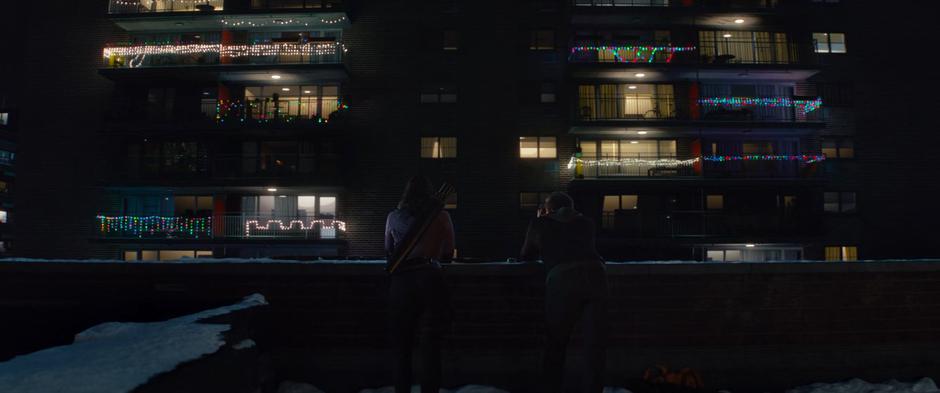 Kate and Clint lean on the edge of the roof across from their target apartment watching.