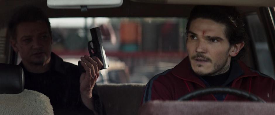 Kazi looks at Clint in the rearview mirror as Clint holds up the gun he stole from the front of the car.