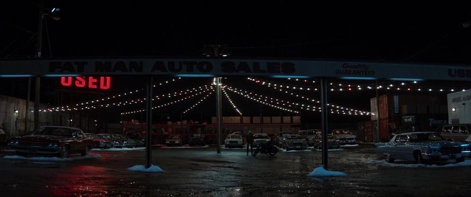 Maya stands next to her motorcycle in the seemingly empty used car lot at night.