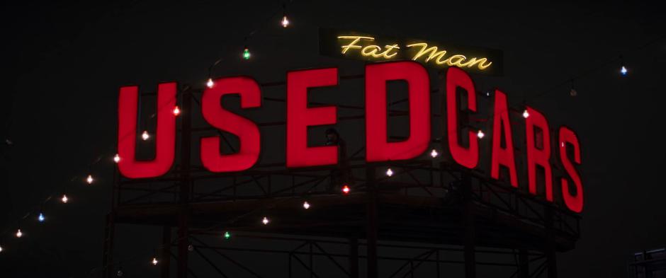 Kazi hides behind the used cars sign with a sniper rifle trained on the lot below.