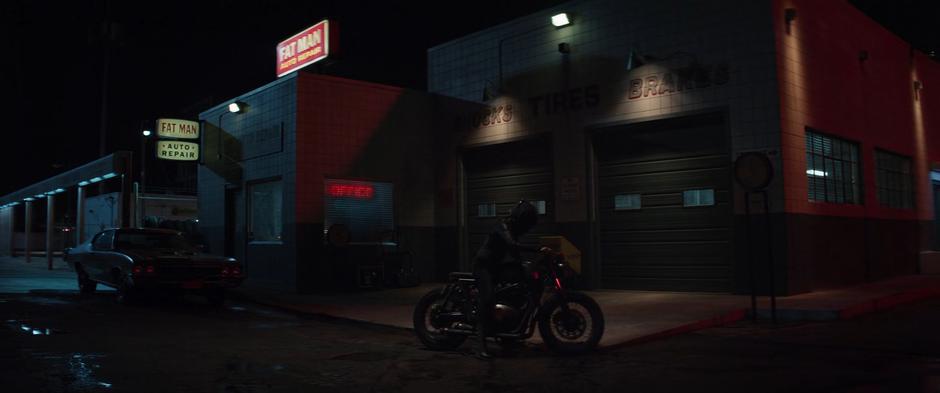 Maya stops her motorcycle in front of hte auto shop at night.