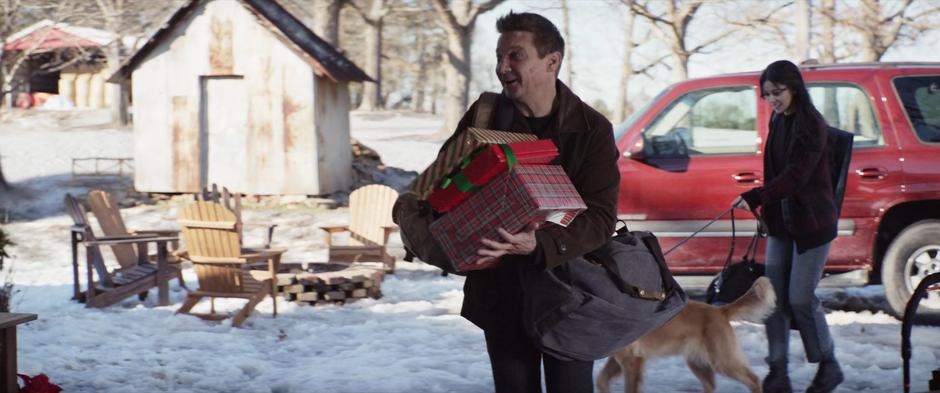 Clint smiles at Nathaniel as he approaches the kid with a handful of presents and Kate walks behind them holding Lucky's leash.