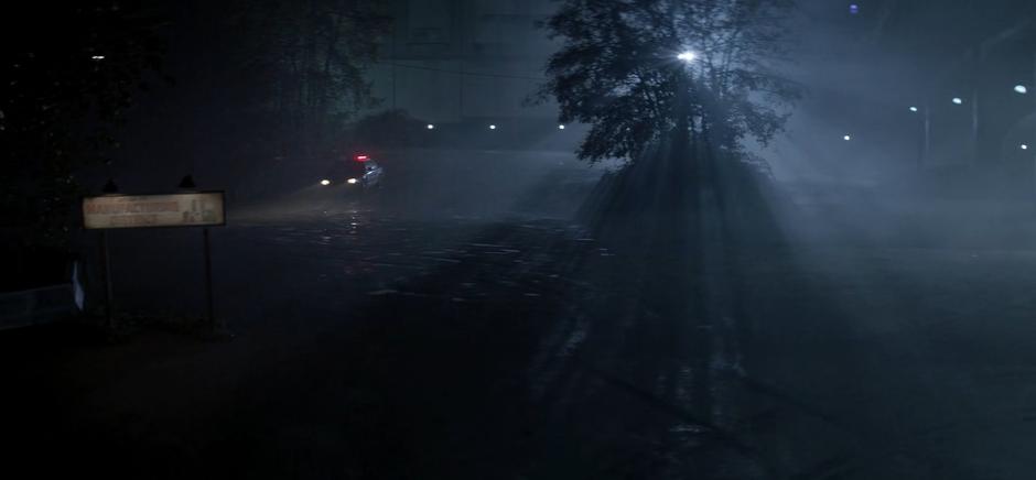 A police car drives through a dirt lot near a factory at night.