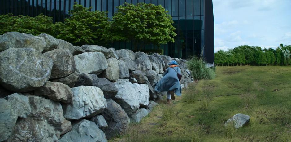 Angela runs hunched over along the rock barrier out of sight of the two goons towards the next building.