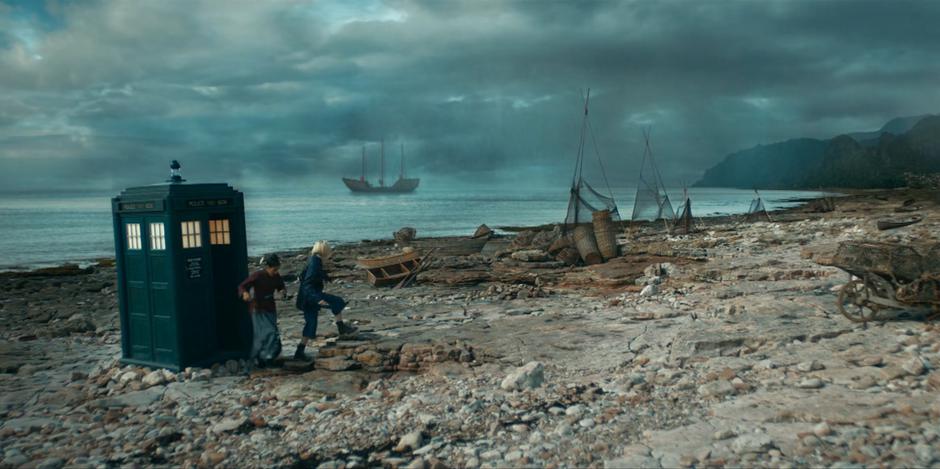 Yaz and the Doctor step out of the TARDIS onto the beach where a ship floats in the distance.