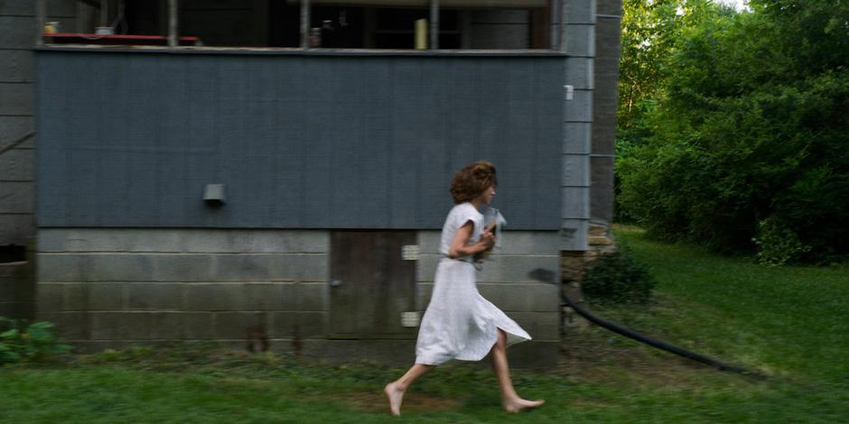 Nancy runs around the side of the house with her bag and shoes in her arms.