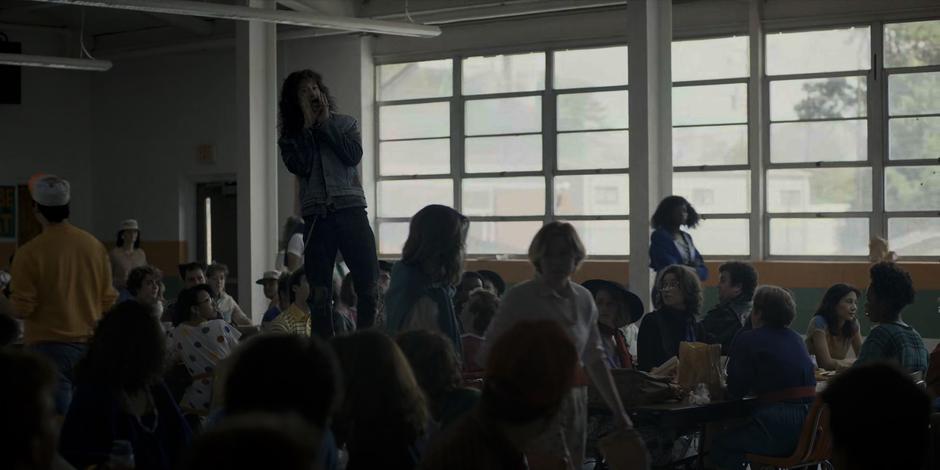 Eddie stands on the table and calls out across the cafeteria.