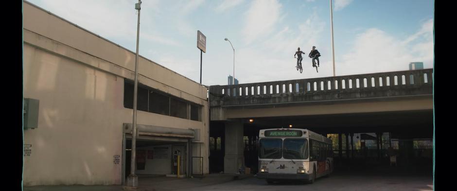 Kamala and Bruno leap their bikes off the bridge over the bus passing below with an Avengercon sign.