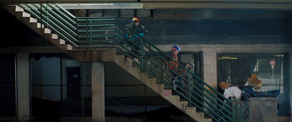 Kamala and Bruno carry their bikes down the stairs towards the bus stop.