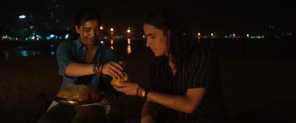 Kamala passes along a bag of biryani to Kareem as they city around the fire.