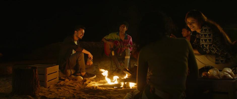 One of Kareem's friends calls out to him while playing a guitar around the fire.
