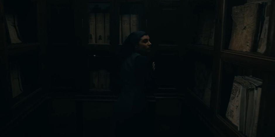 Camila looks over her shoulder as she opens the secret door in a bookshelf.