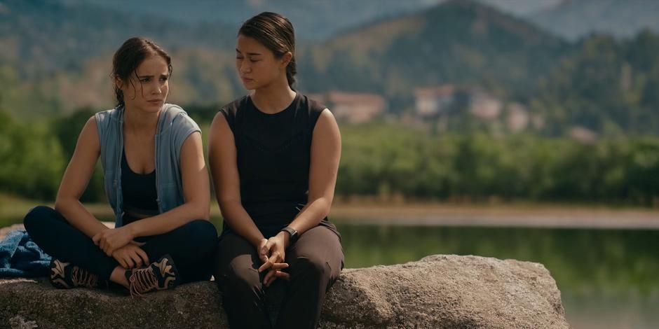 Ava and Bea sit next to each other on the rock in the lake talking.