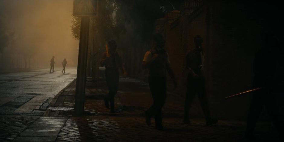 Miguel and Ava cross the street in the distance following several masked figures.