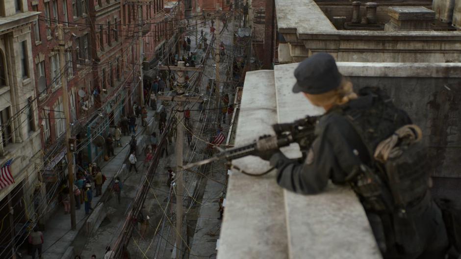 A FEDRA guard watches from a rooftop while people walk the street below.