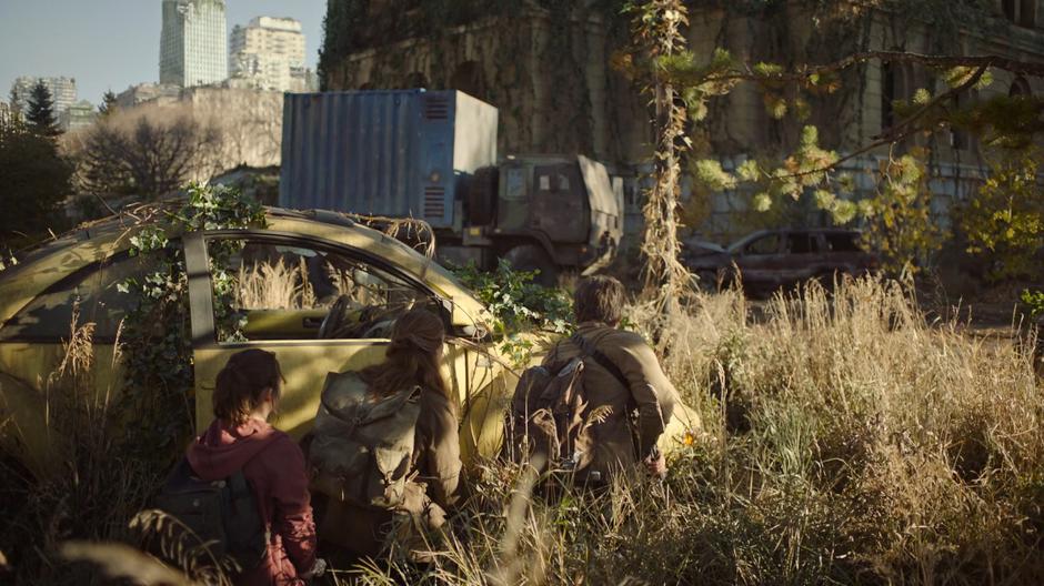 Ellie, Tess, and Joel crouch behind a car as they look for signs of the Fireflies.