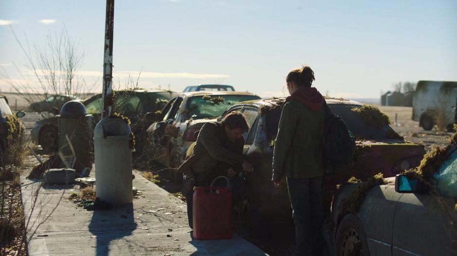 Ellie watches Joel as he siphons gas from a car.