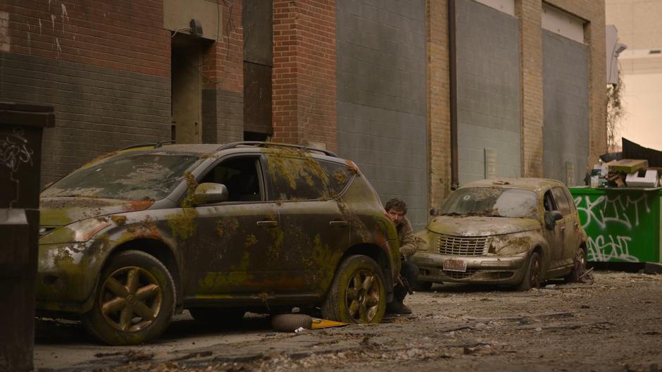 Joel peeks around the side of a ruined car watching a humvee drive past.