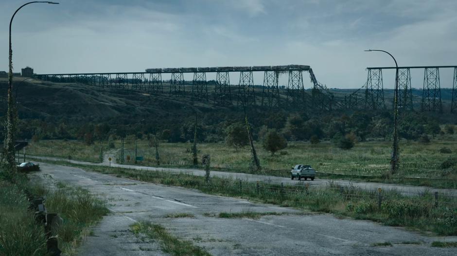 Ellie and Joel drive down the highway as a collapsed railway trestle is visible in the distance.