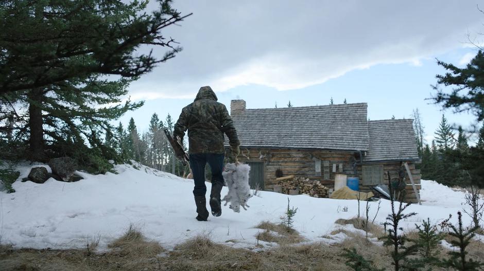 Marlon exits the forest carrying a brace of rabbits and approachs his home.