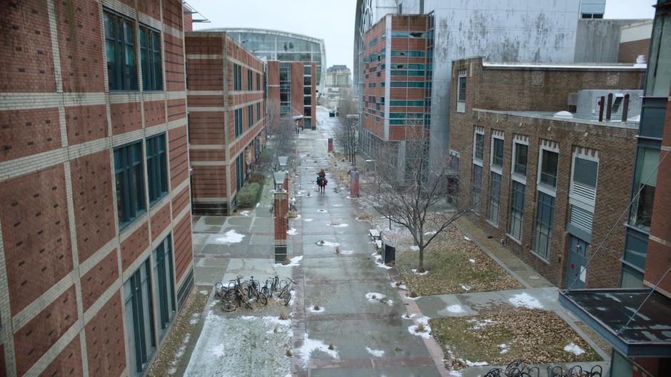 Joel and Ellie ride down a path between two buildings.