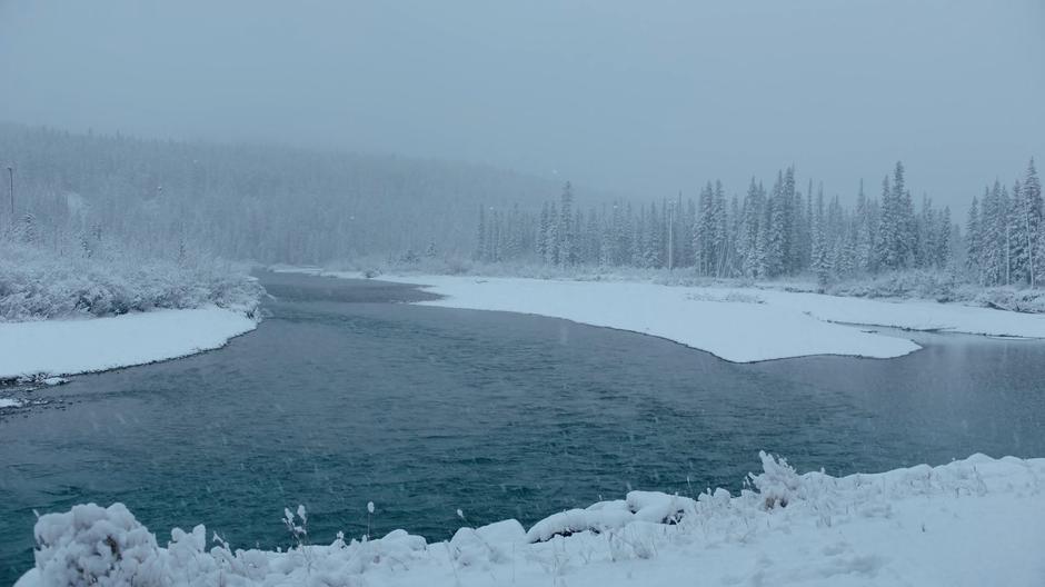 Snow falls on the river surrounded by forests.