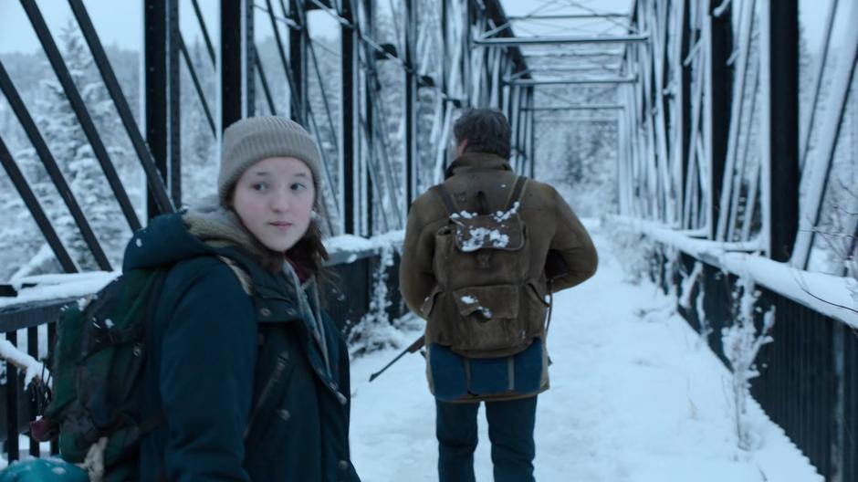 Ellie glances over her shoulder as she follows Joel across the bridge.