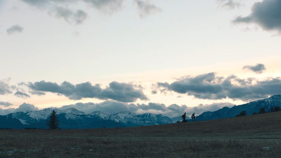 Joel and Ellie climb a hill as the sun disappears behind the mountains.