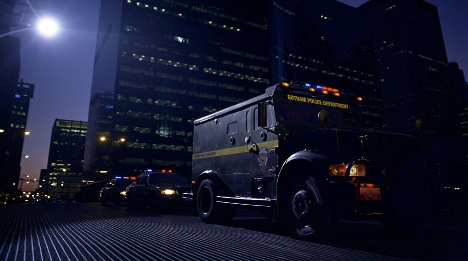 The armored car carrying Harvey Dent drives across a bridge surrounded by a police escort.