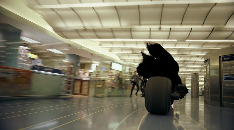 A civilian jumps out of the way as Batman races his Batpod through the shopping center.