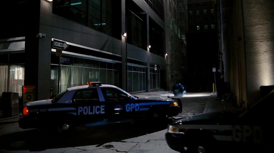 Batman drives the Batpod into the alley while the police cars stop on the street.