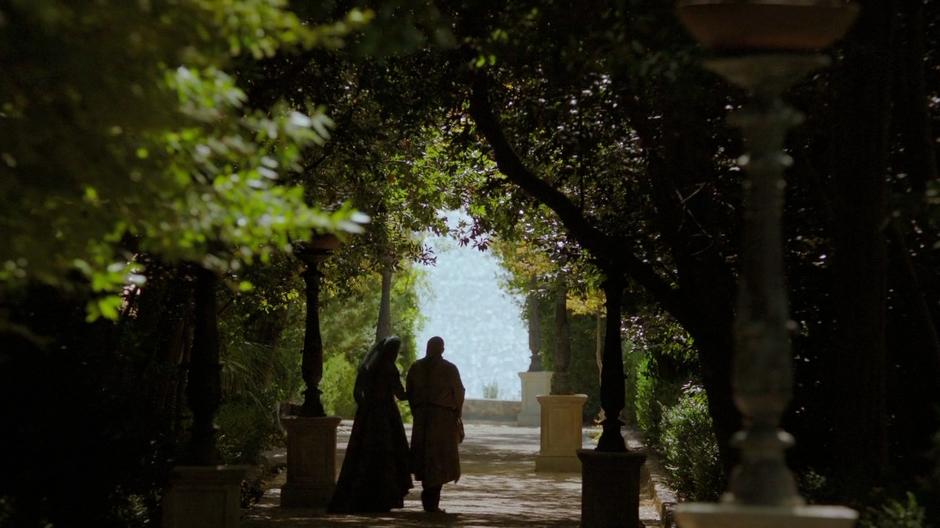 Olenna and Varys talk while walking underneath the trees.