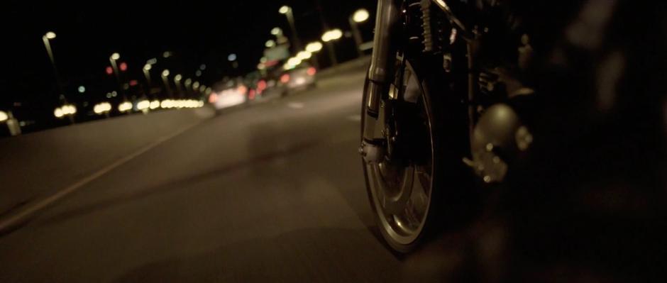 Sam races his motorcycle through traffic over a bridge.