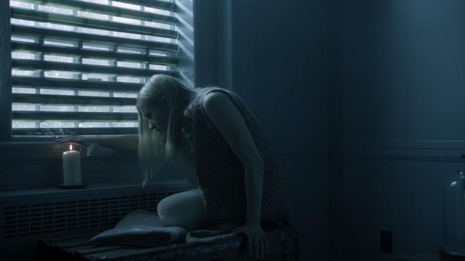 Alice sits by the window in her room holding her hand over a lit candle.