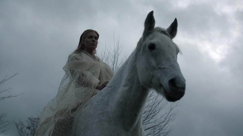 The fairy queen sits on her horse in front of some trees.