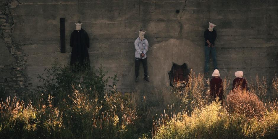 Offred and Ofglen look up at three bodies hanging from the wall.