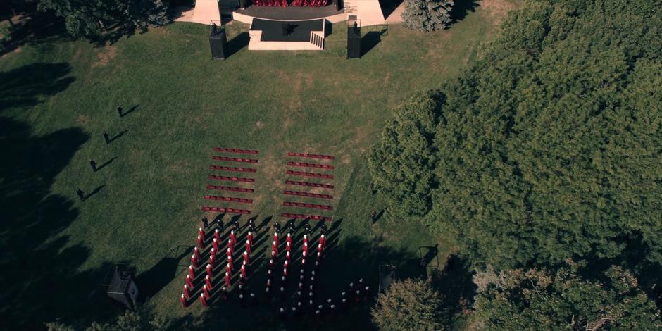 A view from above where the handmaids are assembled into two squares in front of the stage while guards stand in a line off to the side.