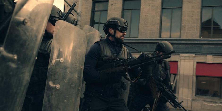 One of the riot offices holds a large machine gun on the crowd while the others line up with their riot shields.