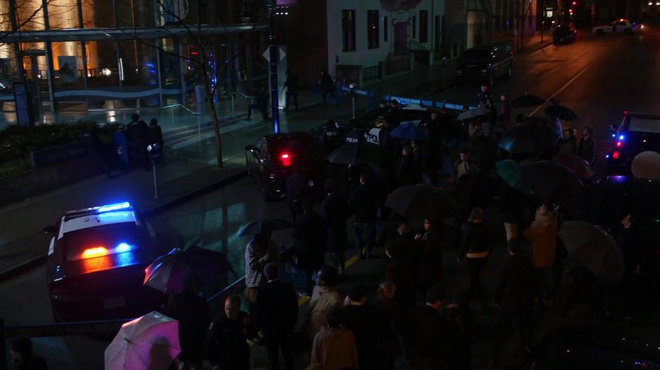 A crowd of onlookers and reporters mills around the street while the police hold a cordon around the bank.