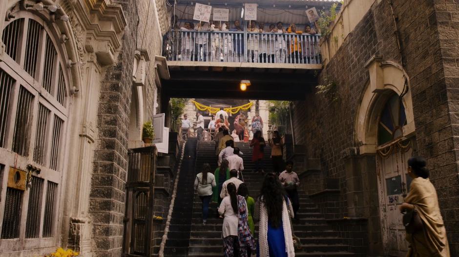 Kala's mother walks towards the front steps while protestors line the overhead walkway.