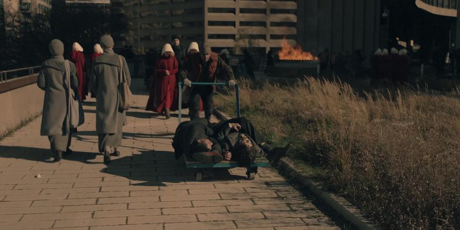 A man pushes a cart of bodies down the path while a bonfire burns in the background.